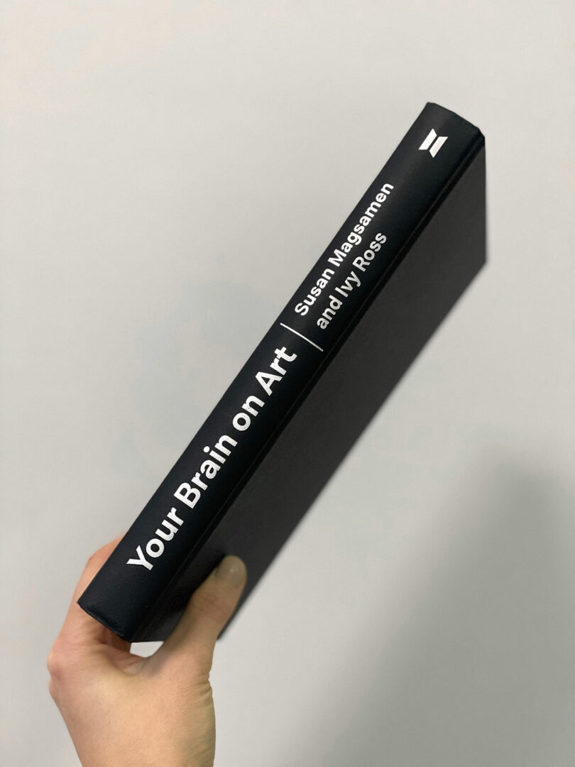 Hand holding a book with a black spine titled "Your Brain on Art" by Susan Magsamen and Ivy Ross against a plain background.