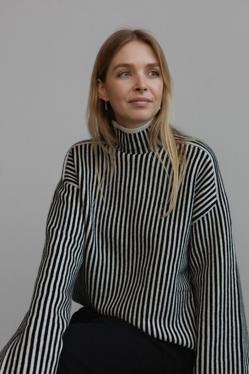 A person with long hair is wearing a black and white striped turtleneck sweater and looking off to the side against a plain background.