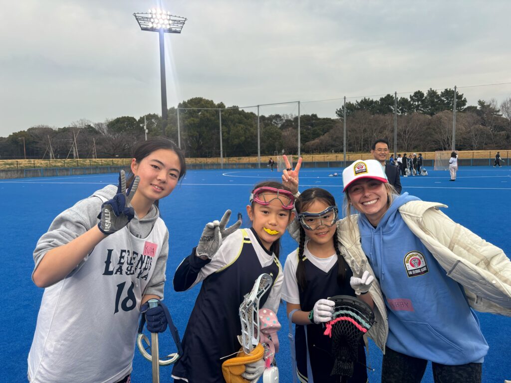 A group of young girls and a woman pose on a blue sports field, smiling and making peace signs. They are dressed in athletic gear.