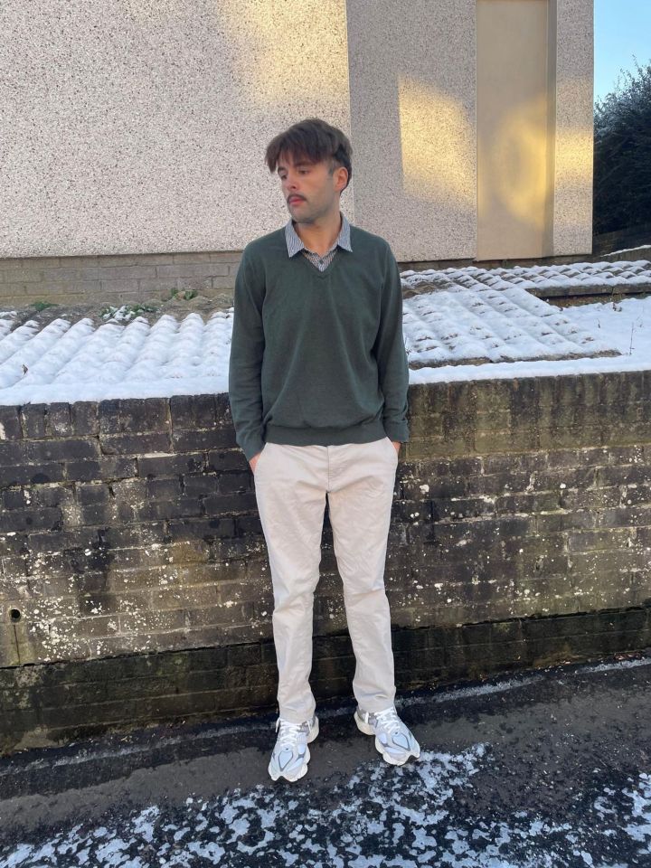 A man in a green sweater and beige pants stands against a brick wall.