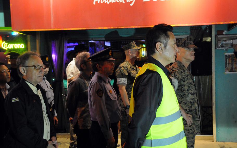 A group of men, including military personnel in camouflage uniforms, police officers and civilians, stand in formation on a sidewalk at night in front of a brightly lit bar with a red sign. One man in the foreground wears a reflective safety vest. The atmosphere suggests a coordinated event or patrol, possibly related to community safety or a formal inspection. 