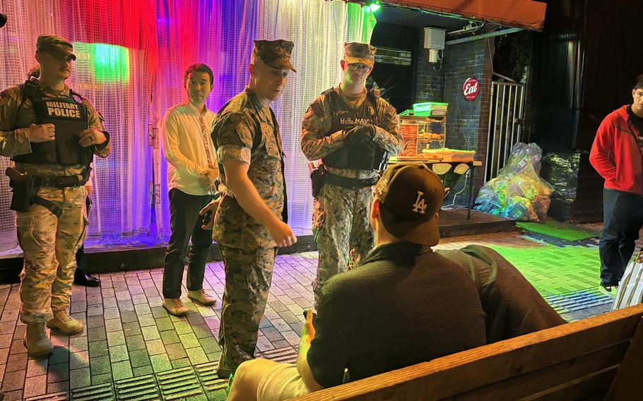 Military police and  Col. David Banning interact with service members on a  street at night, near a colorful curtain backdrop.