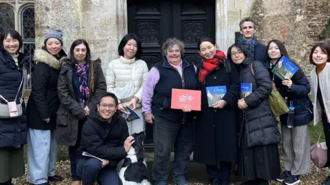 BBC A group of eight Japanese tour operators and two representatives from Visit Britain pose with Caroline Lowsley-Williams, owner of Chavenage House, in the middle. Most are smiling at the camera and some are holding up a Chavenage House brochure. They are stood outside a large ornate black door, and the mottled Cotswold stone walls of the house can be seen behind them. 
