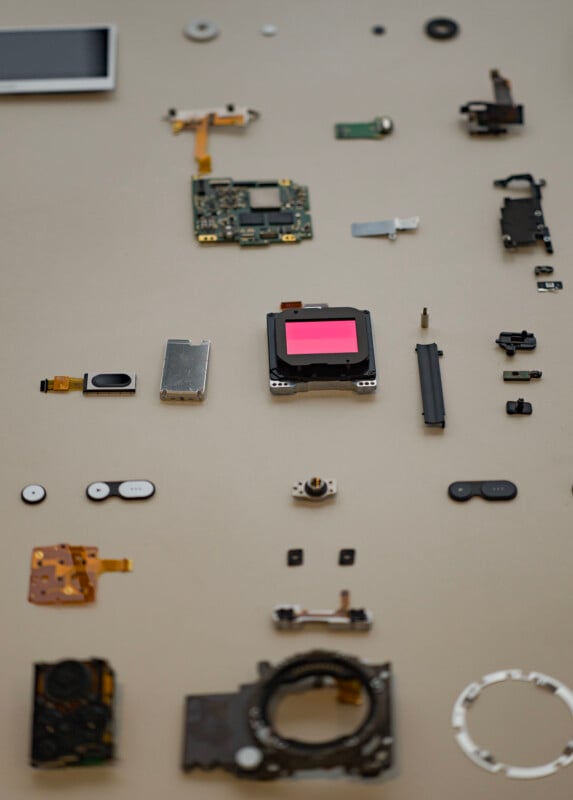 Various electronic components are laid out on a beige surface, including circuit boards, connectors, and metallic parts. The components are arranged in a grid-like pattern, showcasing the intricate details and variety of pieces used in electronic assembly.