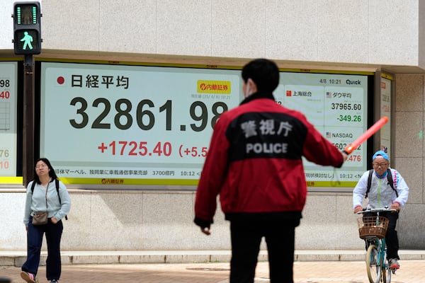 Monitors, back, show Japan's Nikkei 225 index at a securities firm as a member of the Tokyo Metropolitan Police directs traffic and pedestrians for intersection safety in the morning in Tokyo, Tuesday, April 8, 2025. (AP Photo/Hiro Komae)