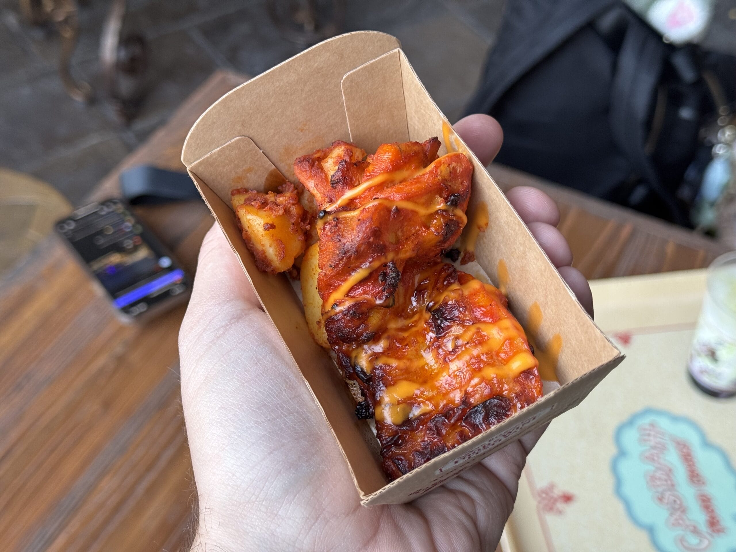 A hand holds a cardboard tray of cheesy, baked chicken bites at the 2025 Food and Wine Festival; phone in background.