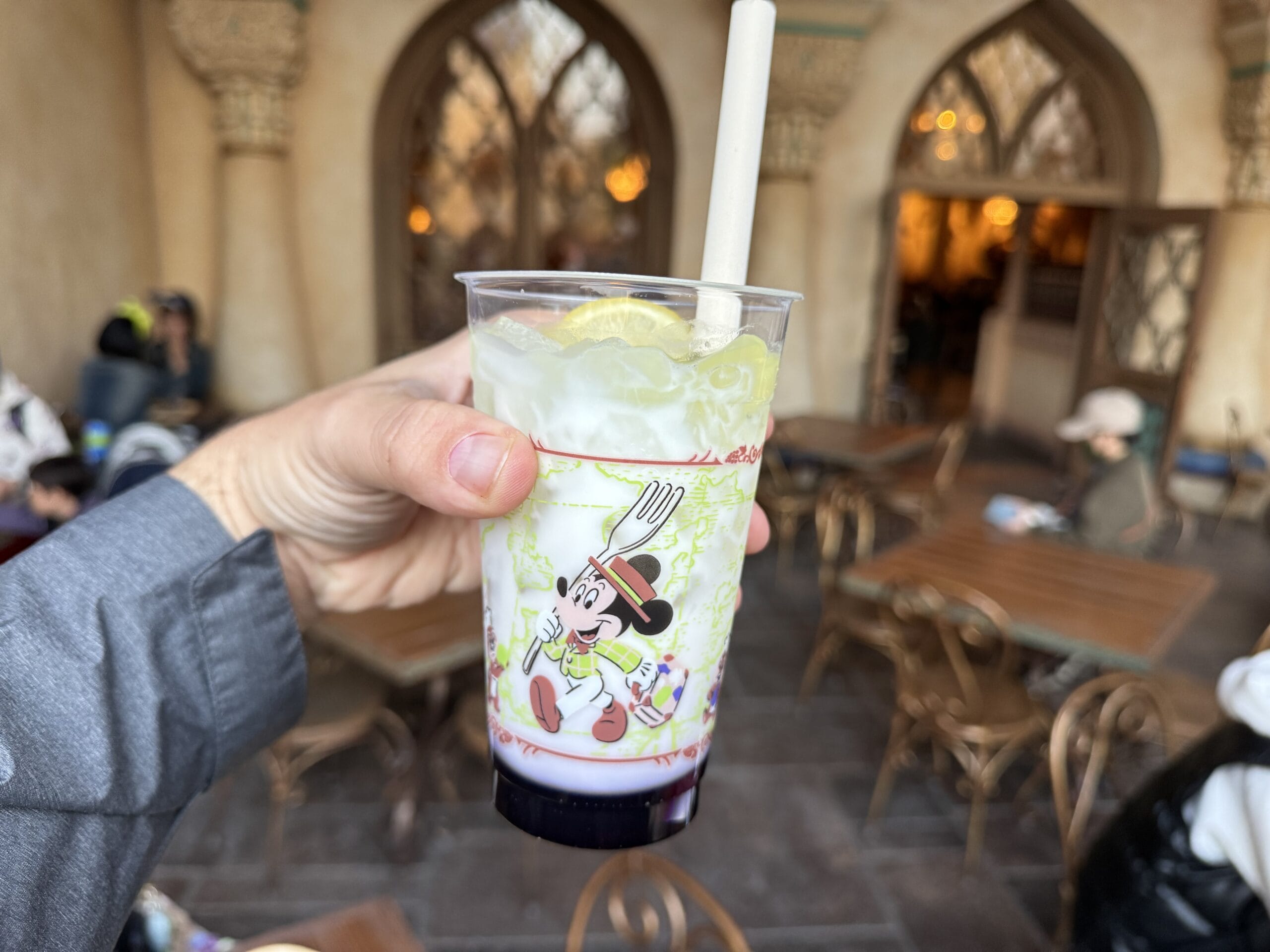 A hand holds a cold drink with lemon in a Chef Mickey cup at Tokyo DisneySea’s outdoor café during the 2025 Food and Wine Festival.