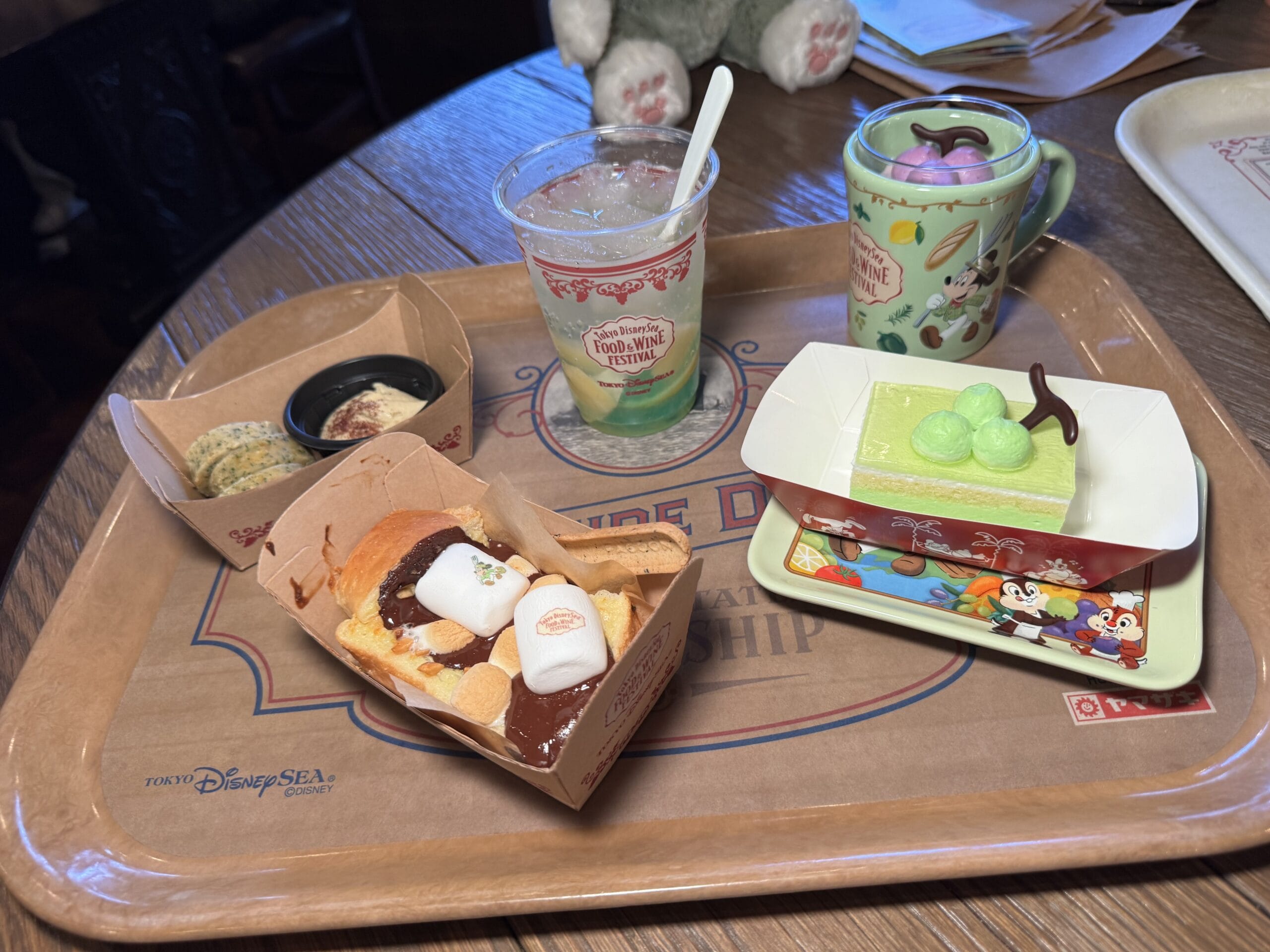 A tray with a dessert pastry topped with marshmallows, a green parfait, a slice of green cake, a cup with a drink, and cookies—delights served at the 2025 Food and Wine Festival in Tokyo DisneySea.