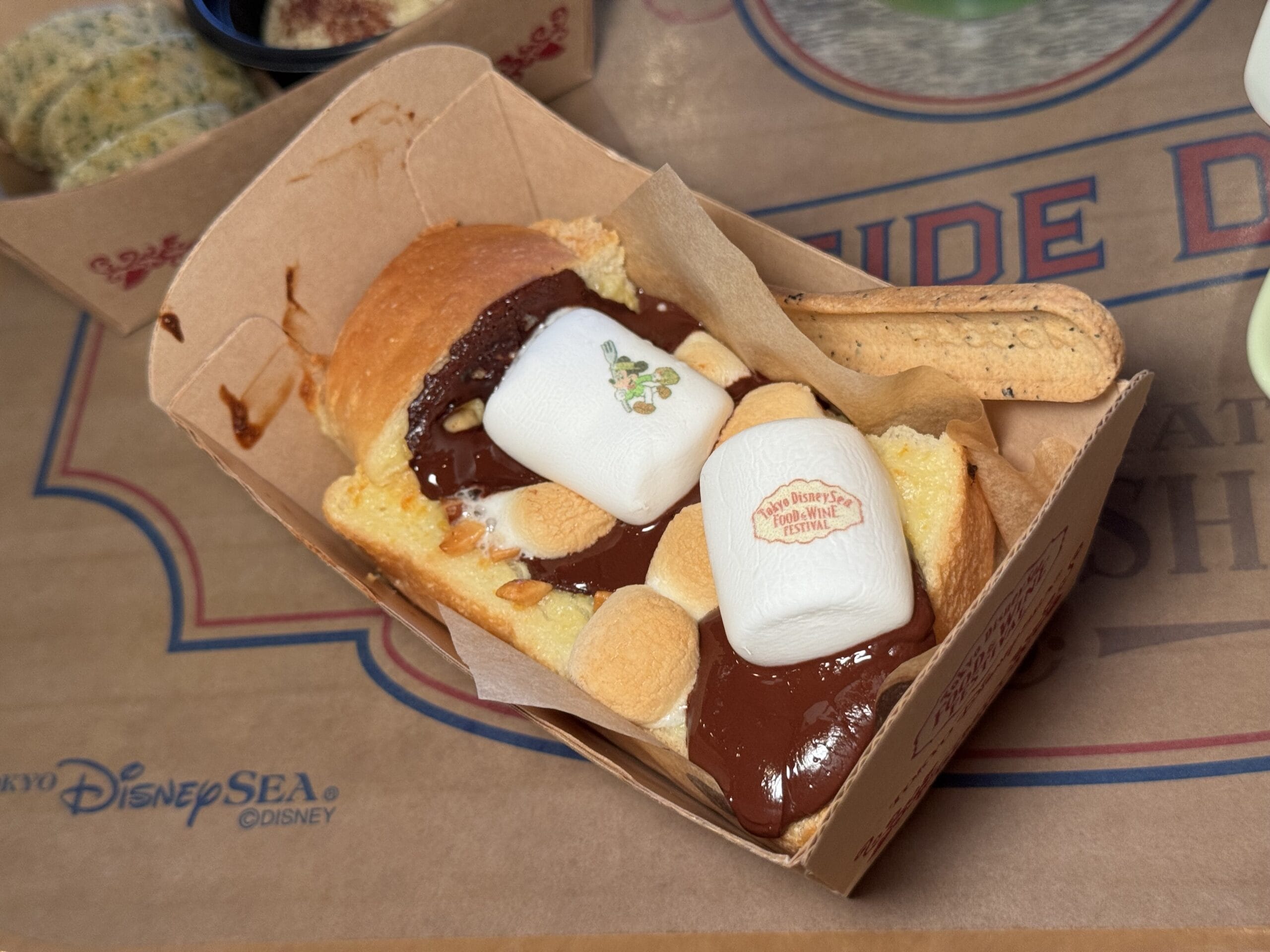 A rectangular cardboard tray holds bread topped with melted chocolate, marshmallows, and a biscuit, served on Tokyo DisneySea-themed paper—perfect for a sweet treat at the 2025 Food and Wine Festival.