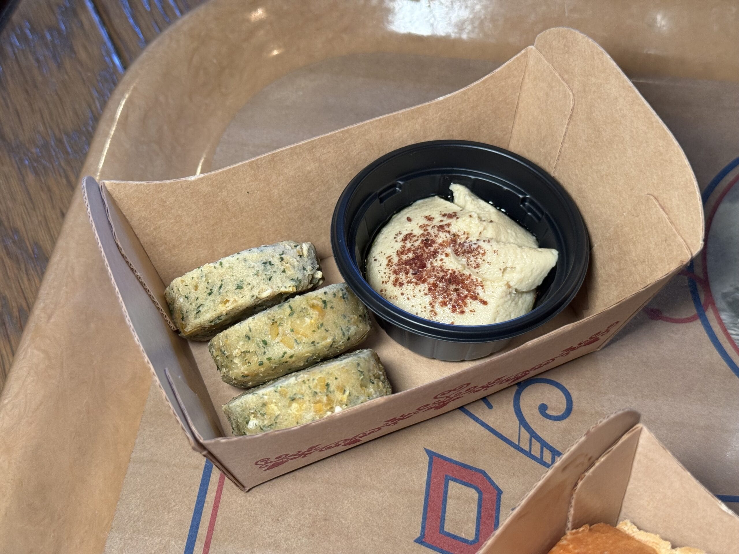 Three pieces of falafel and a small container of hummus with spice garnish, served in a cardboard tray—perfect for enjoying at the 2025 Tokyo DisneySea Food and Wine Festival.