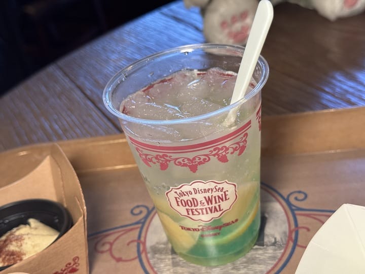 A plastic cup with a lemon-lime drink, ice, and a white straw sits on a tray. The cup displays the "Tokyo DisneySea 2025 Food & Wine Festival" logo.