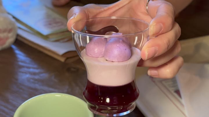 At the 2025 Food and Wine Festival, a hand holds a glass cup filled with layered purple and white mousse topped with two dollops of purple cream; a cup and papers are on the table nearby.