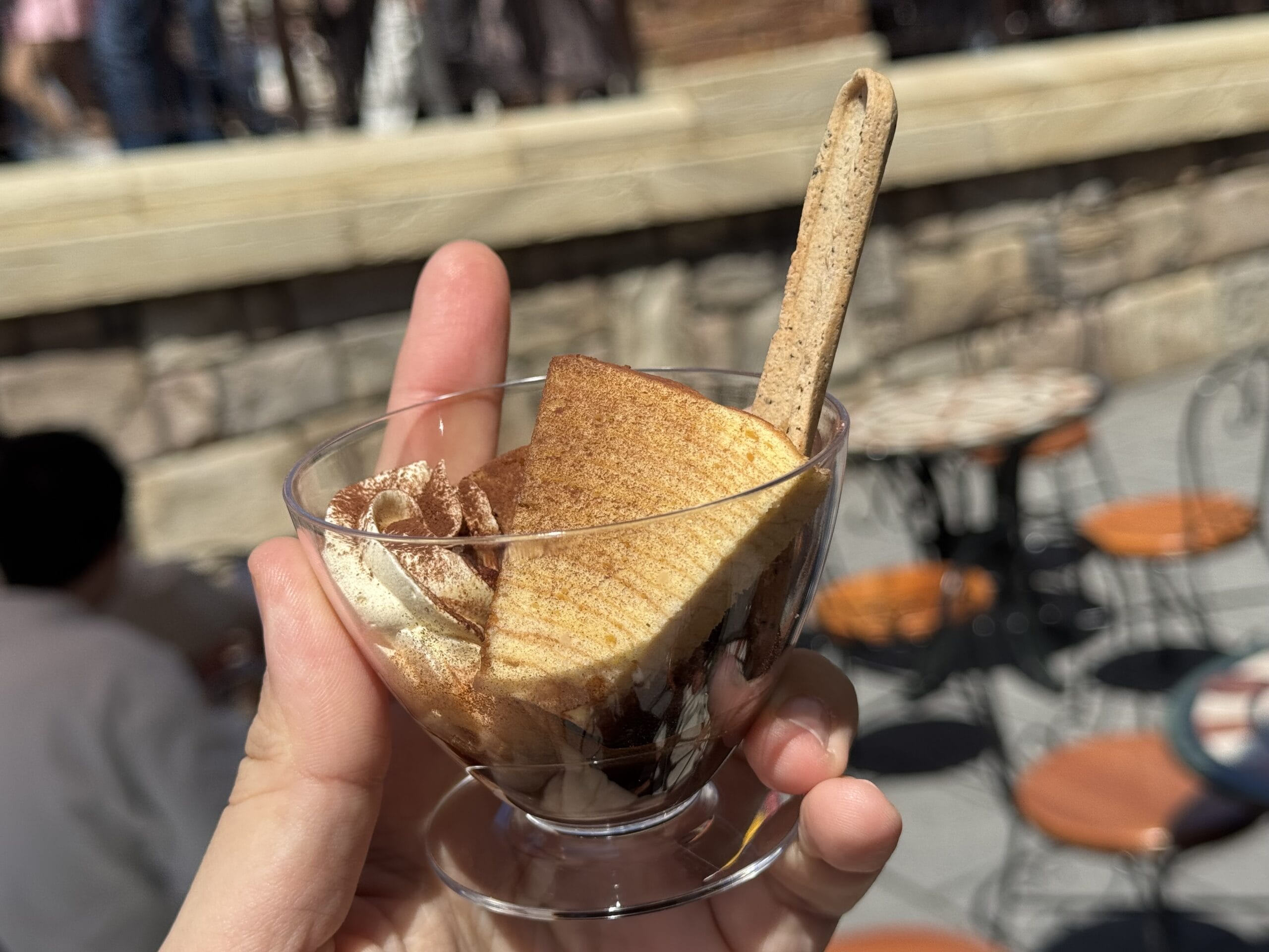 A hand holds a glass cup of tiramisu with a biscuit at Tokyo DisneySea’s 2025 Food and Wine Festival, outdoor seating behind.