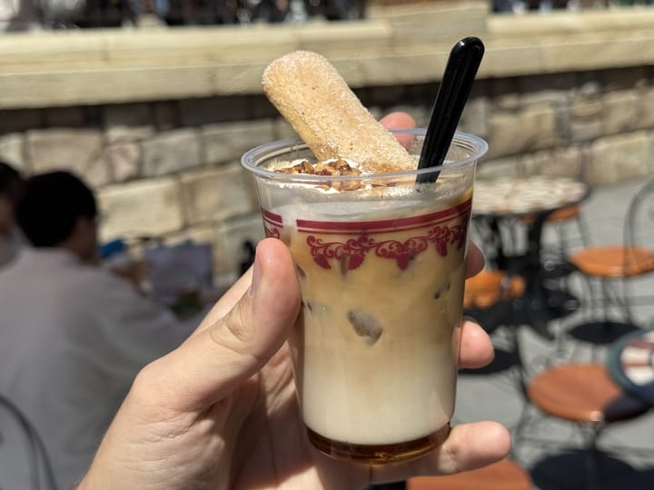 A hand holds layered iced coffee with a ladyfinger, at Tokyo DisneySea's 2025 Food and Wine Festival café terrace.