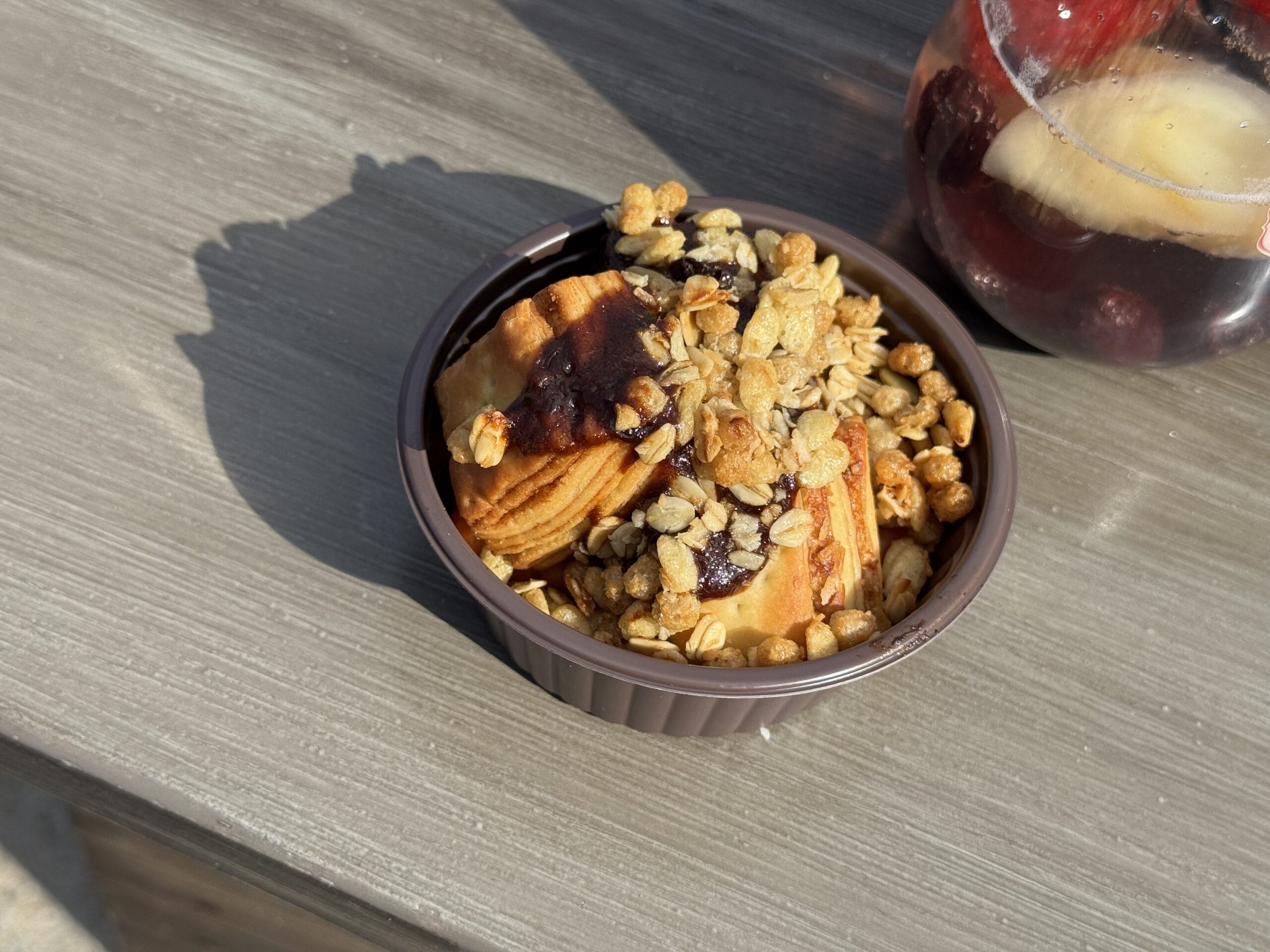 A small brown bowl filled with pastries topped with granola and dark syrup sits on a wooden surface at the Tokyo DisneySea Food and Wine Festival 2025; a glass with fruit is partially visible in the background.