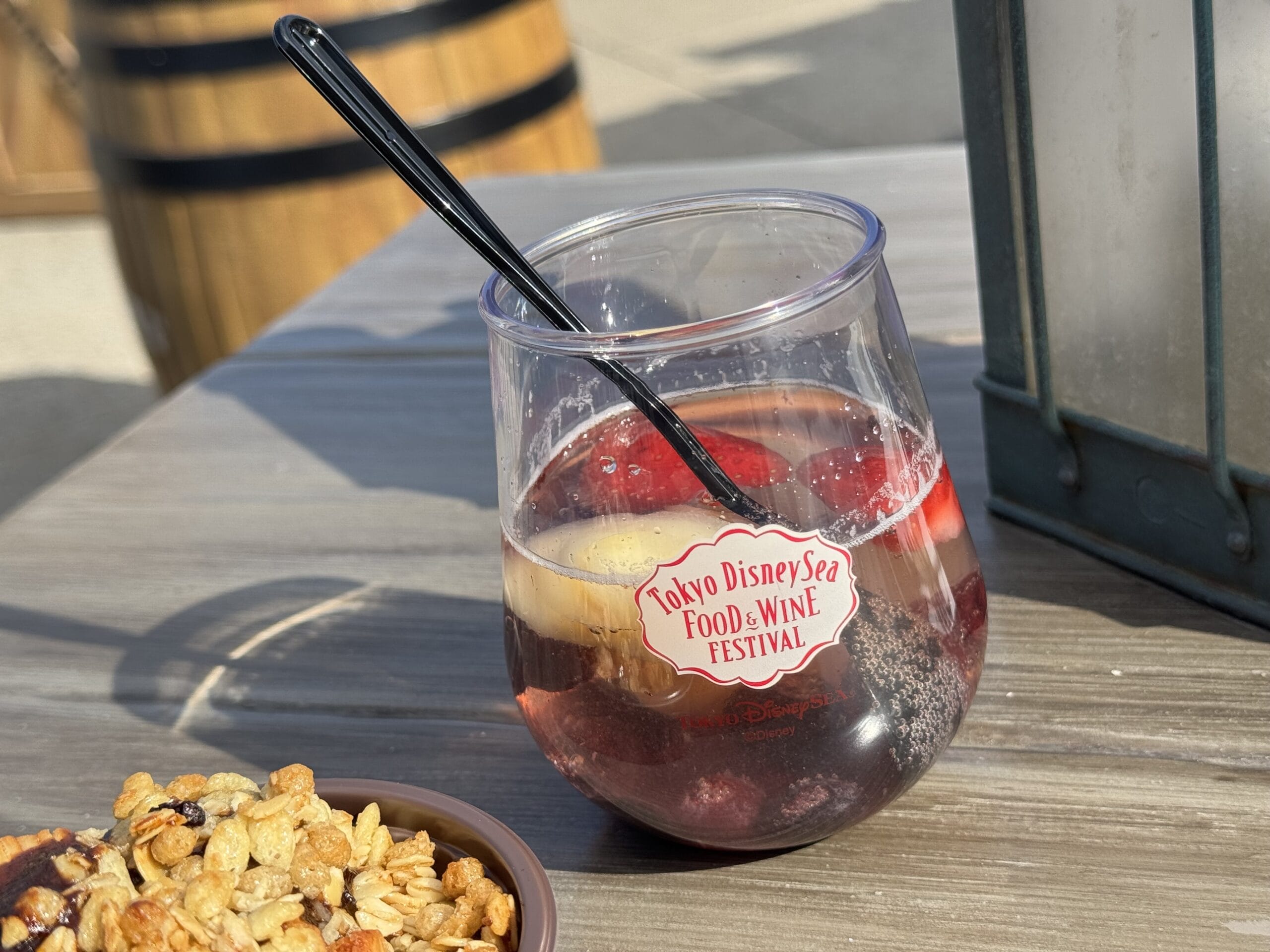 A clear plastic cup with a “Tokyo DisneySea Food and Wine Festival 2025” logo holds a berry fruit drink with a black straw, set on a wooden table beside a bowl of granola.