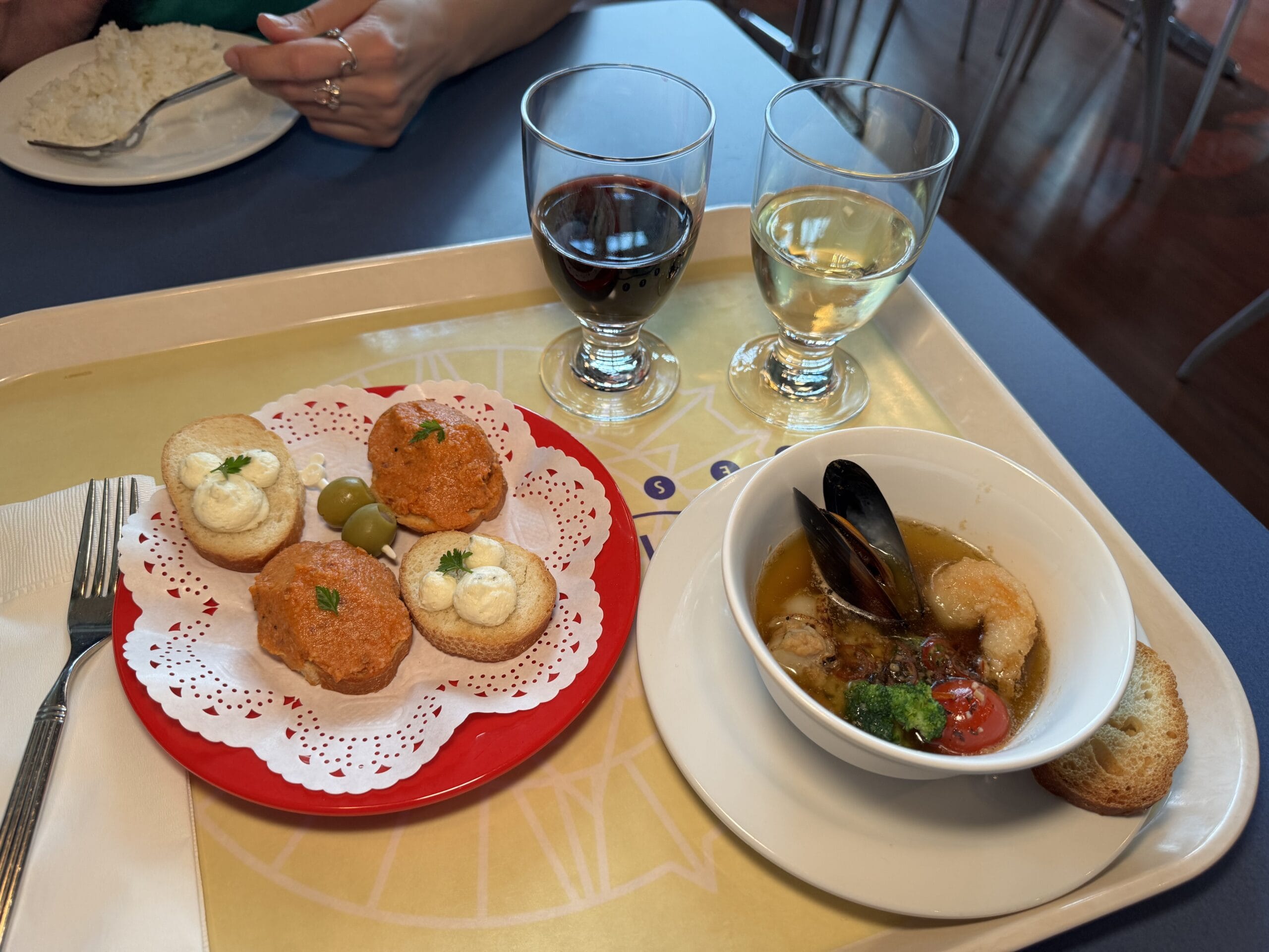 A tray at the 2025 Tokyo DisneySea Food and Wine Festival features two glasses of wine, a bowl of seafood soup with bread, and a plate of savory mousse appetizers garnished with herbs and olives.
