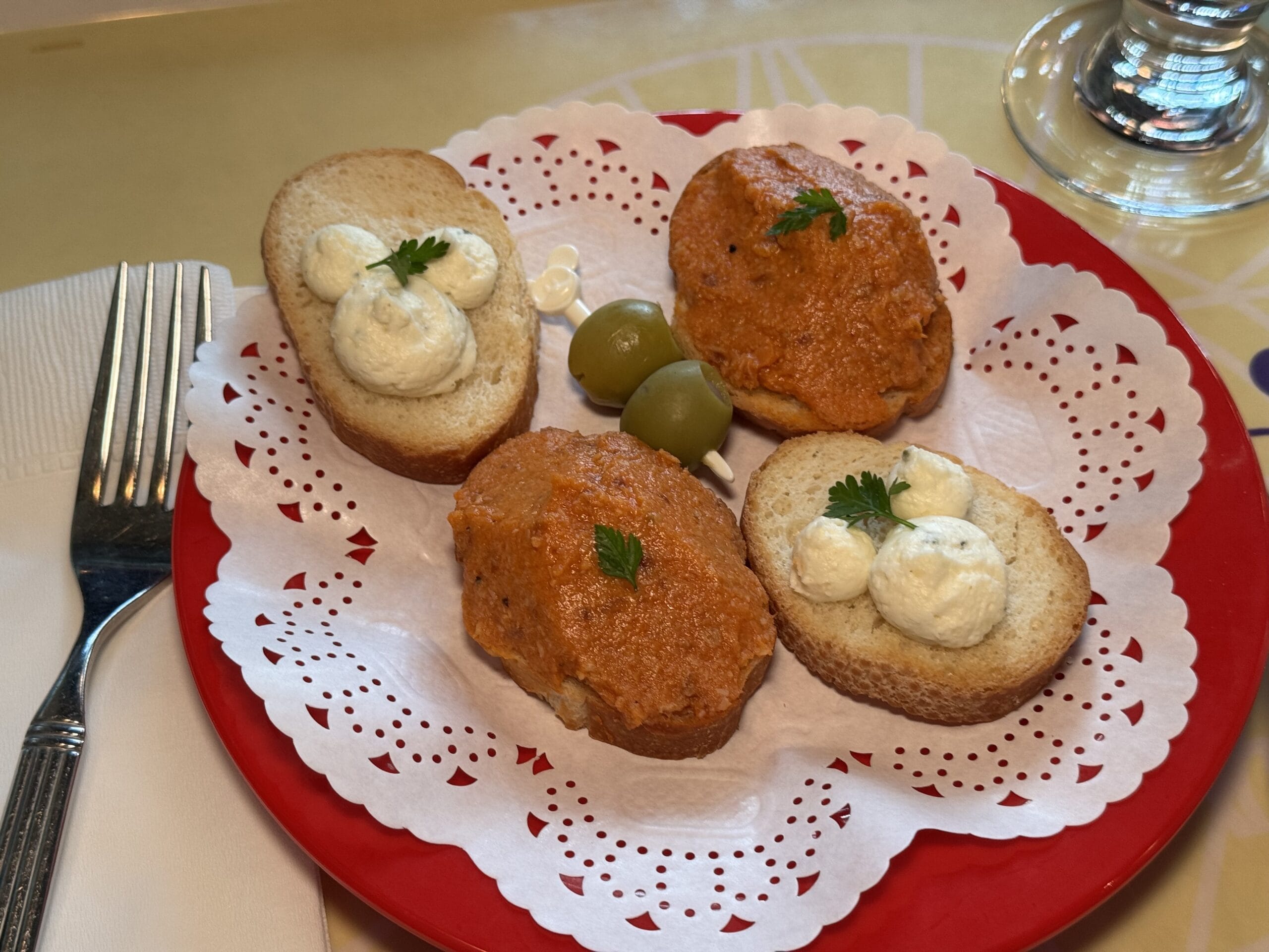 Three slices of bread on a red plate, two topped with white cream and one with a reddish spread; two green olives in the center, garnished with parsley—perfect for savoring at the 2025 Food and Wine Festival at Tokyo DisneySea. Napkin and utensils beside.