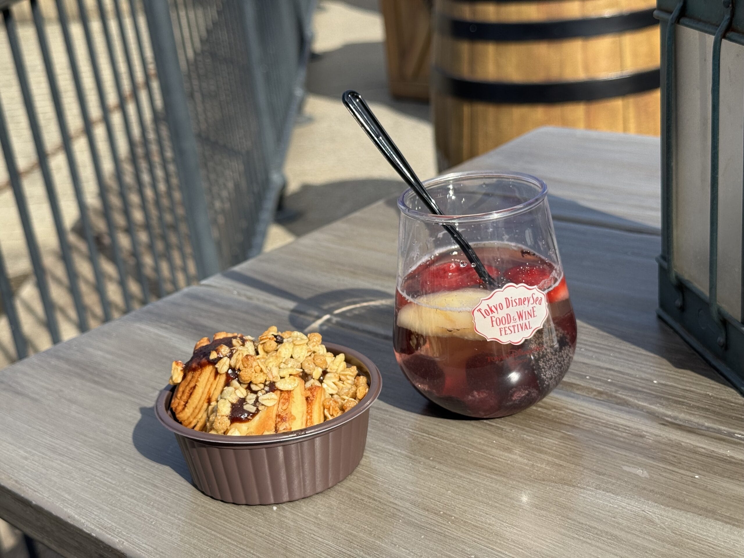 A bowl of pastries topped with granola and chocolate chips sits next to a clear cup of red beverage with ice on a wooden table outdoors, capturing the lively spirit of the 2025 Food and Wine Festival.