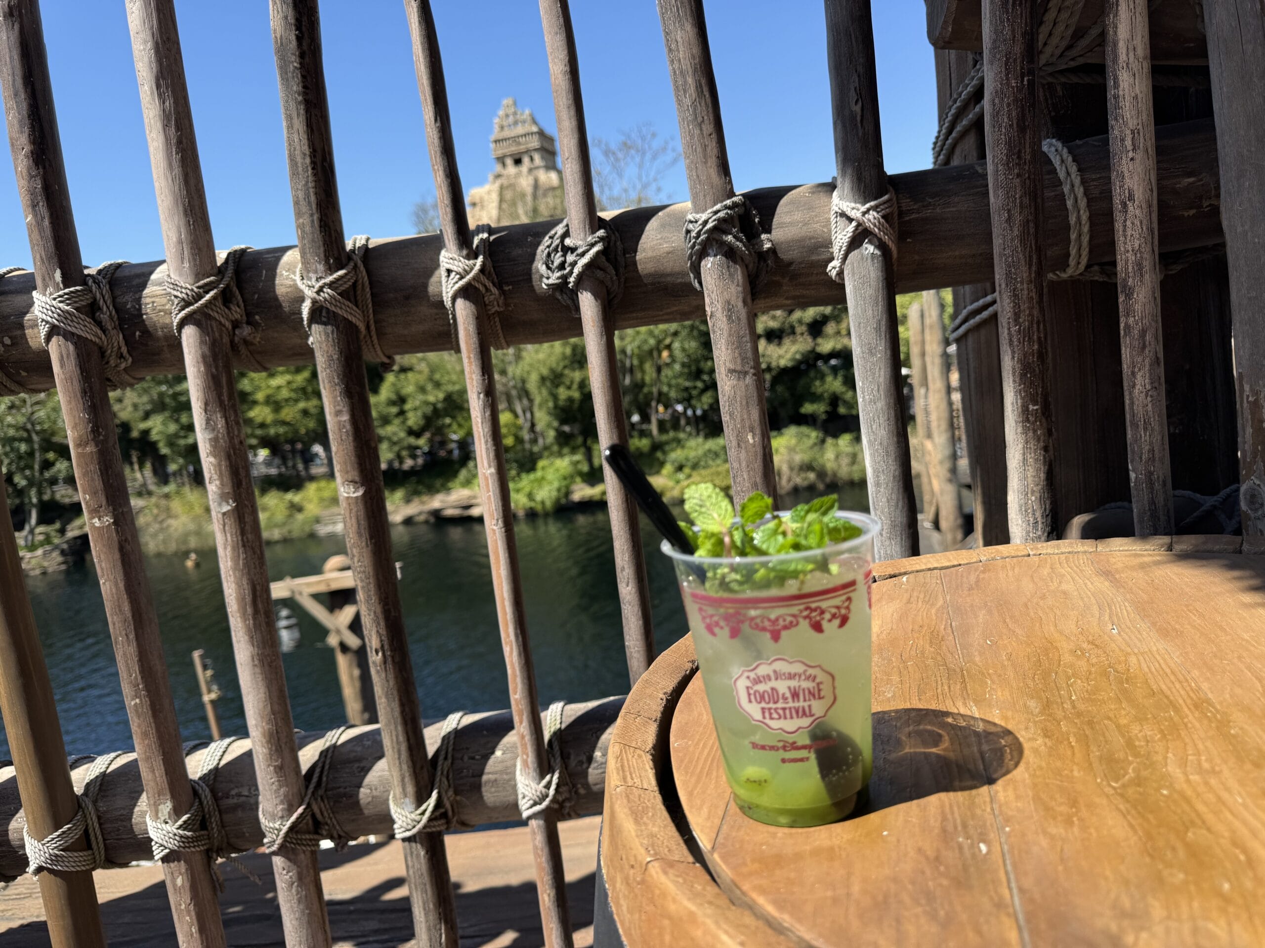 A mint cocktail sits by a bamboo fence at Tokyo DisneySea’s Food and Wine Festival, water and trees in the background.