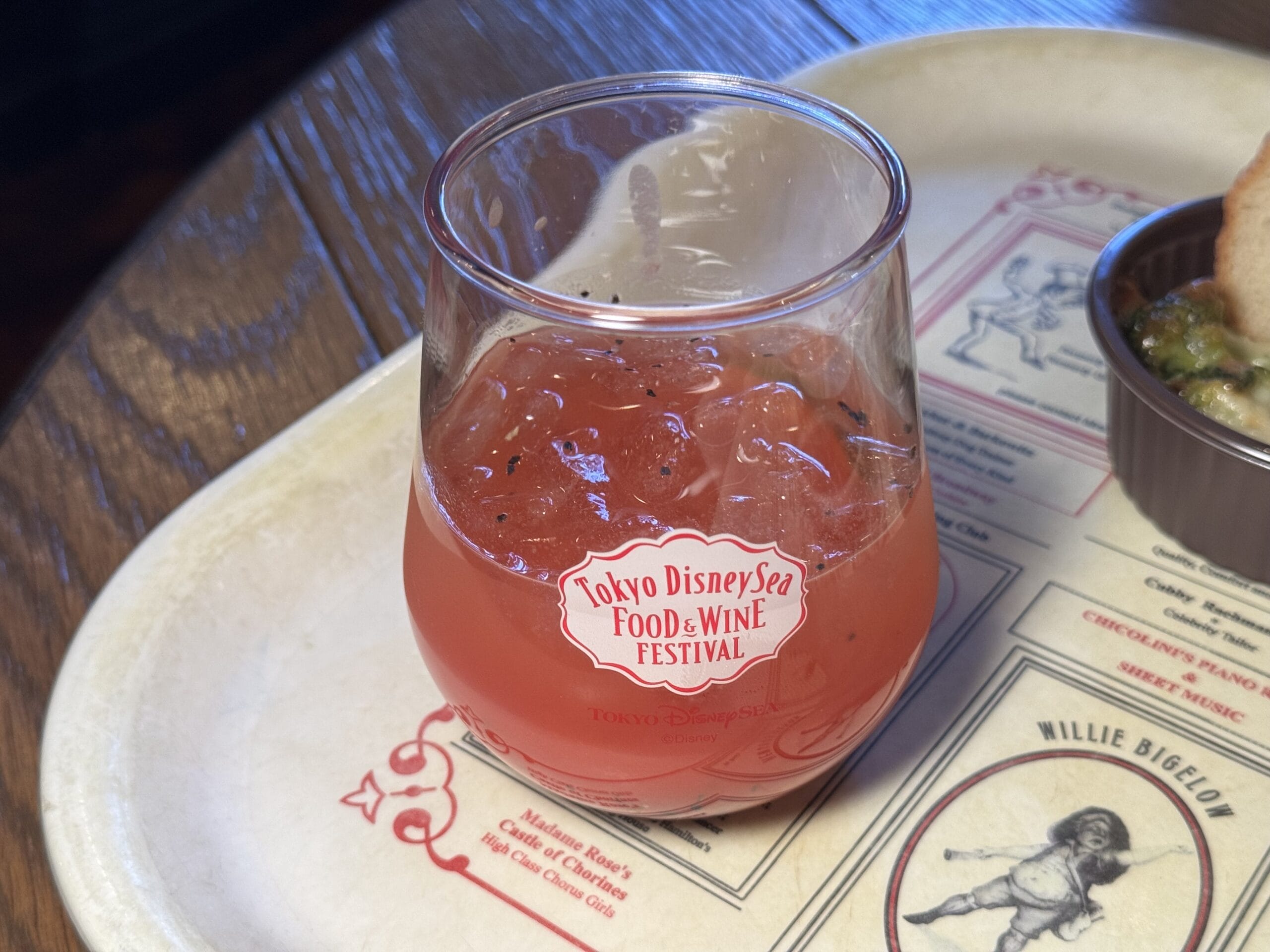 A glass of pink, iced beverage with "Tokyo DisneySea Food & Wine Festival" printed on it sits on a tray with a menu design, capturing the vibrant spirit of the 2025 Food and Wine Festival.