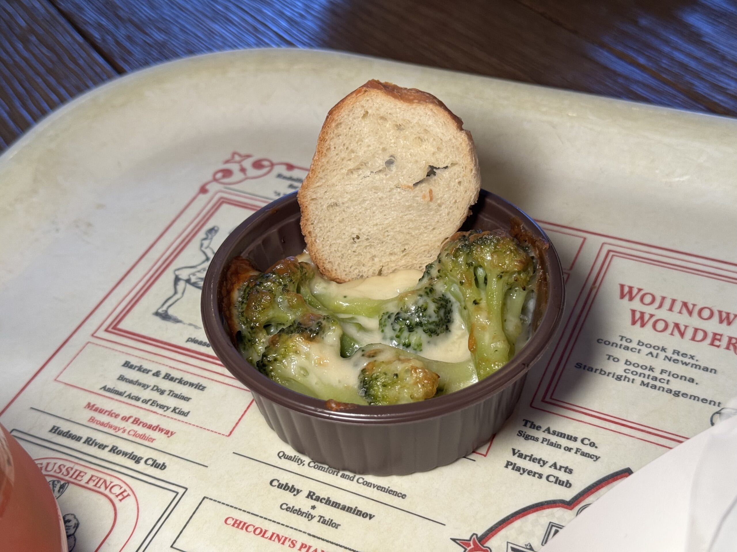 A small brown dish of baked broccoli topped with melted cheese and served with a round slice of toasted bread, placed on a decorative tray—perfect for enjoying at the 2025 Food and Wine Festival.