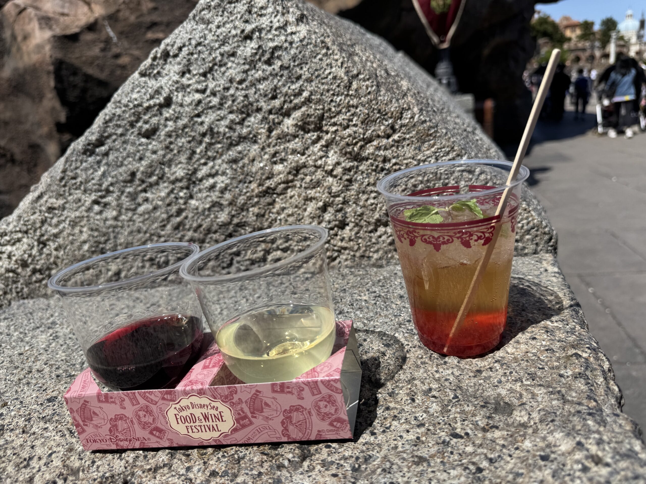 Three drink samples—two in clear cups on a "Food & Wine Festival" tray, and one mixed drink with a stirrer—are placed on a stone surface outdoors at Tokyo DisneySea, perfect for enjoying the festival's flavors in 2025.