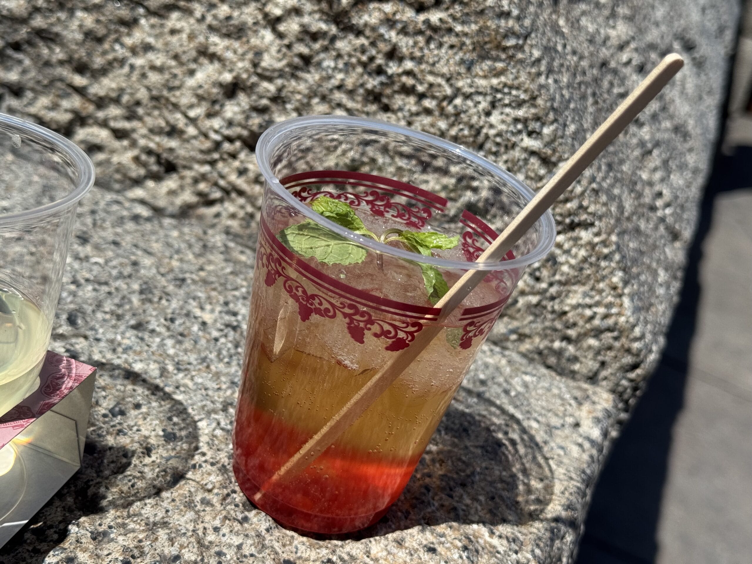A plastic cup with a layered iced drink, garnished with mint leaves and a straw, sits on a rough stone surface in sunlight—perfect for enjoying at the 2025 Food and Wine Festival at Tokyo DisneySea.