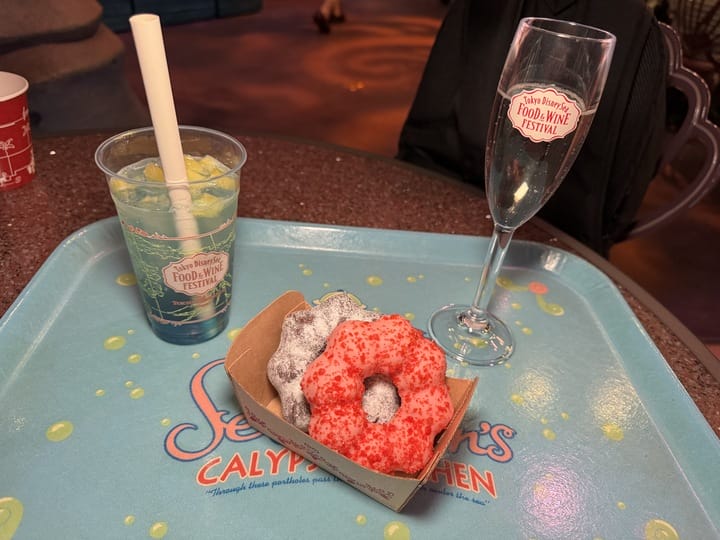 At the 2025 Food and Wine Festival at Tokyo DisneySea, a tray holds a blue drink with a straw, a champagne flute, and two donuts topped with red and white powdered sugar.