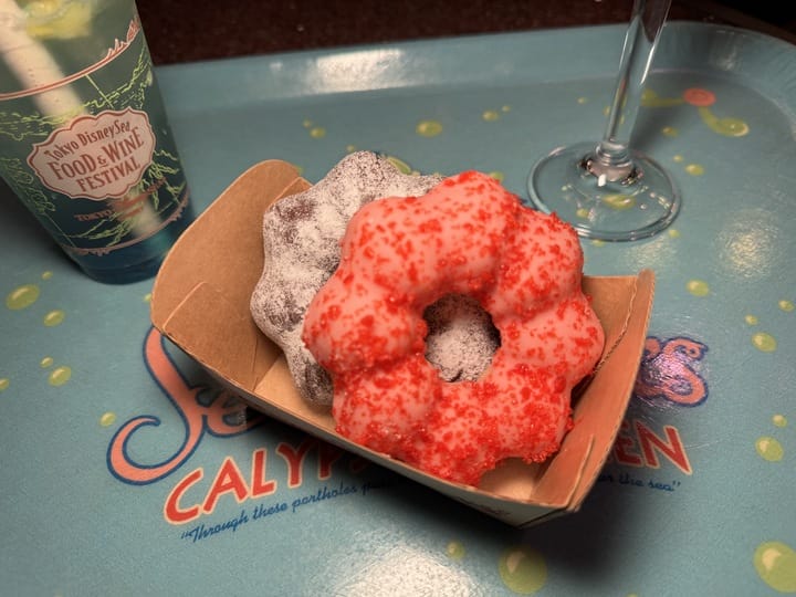 Two bundt-shaped donuts, one dusted with powdered sugar and the other with red sprinkles, sit in a paper tray beside a drink at the 2025 Food and Wine Festival at Tokyo DisneySea.