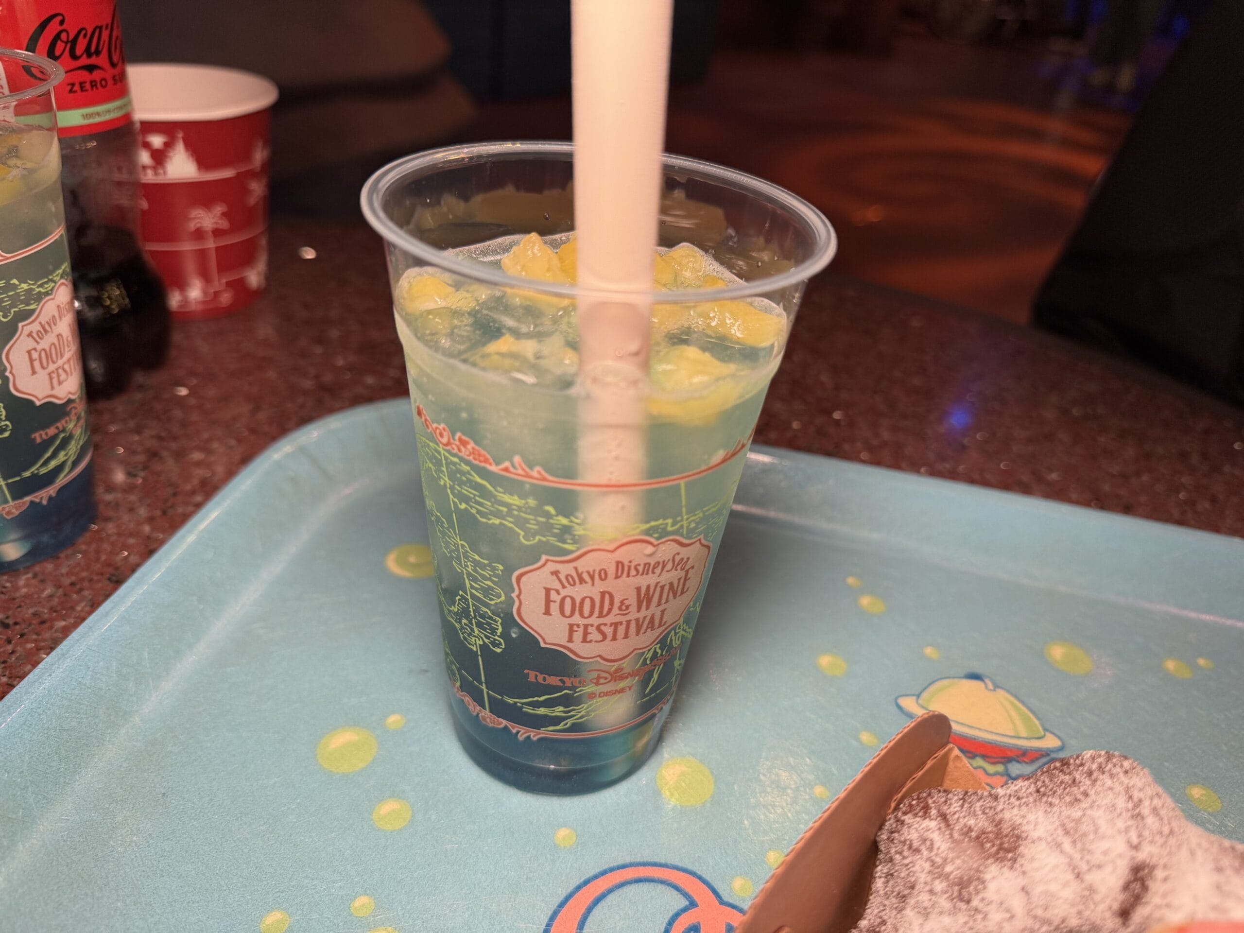 A plastic cup with a light blue and yellow drink and ice sits on a blue tray labeled “Tokyo DisneySea 2025 Food and Wine Festival.” A powdered snack and red cup are partially visible.