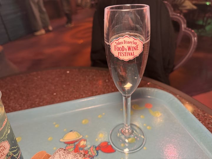 A clear glass with a "Tokyo DisneySea Food and Wine Festival 2025" label sits on a blue tray featuring cartoon sea creatures.
