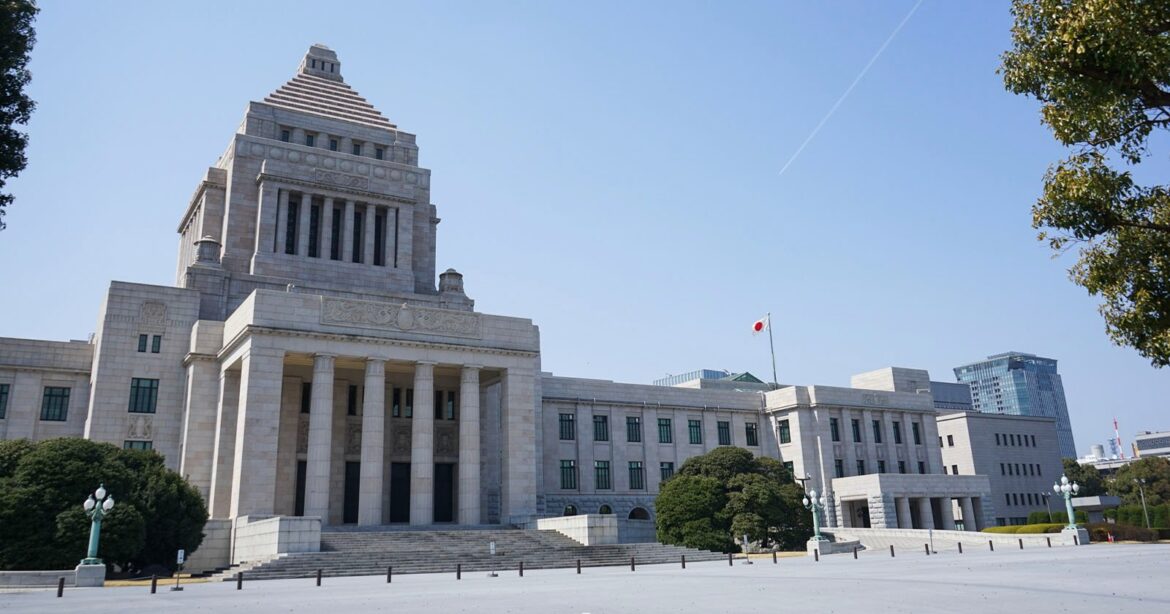 Japan Considers Making AI-Generated Stuio Ghibli Images Illegal A large stone government building with a central pyramid-shaped tower, columns at the entrance, and a Japanese flag on a flagpole, surrounded by trees under a clear blue sky.