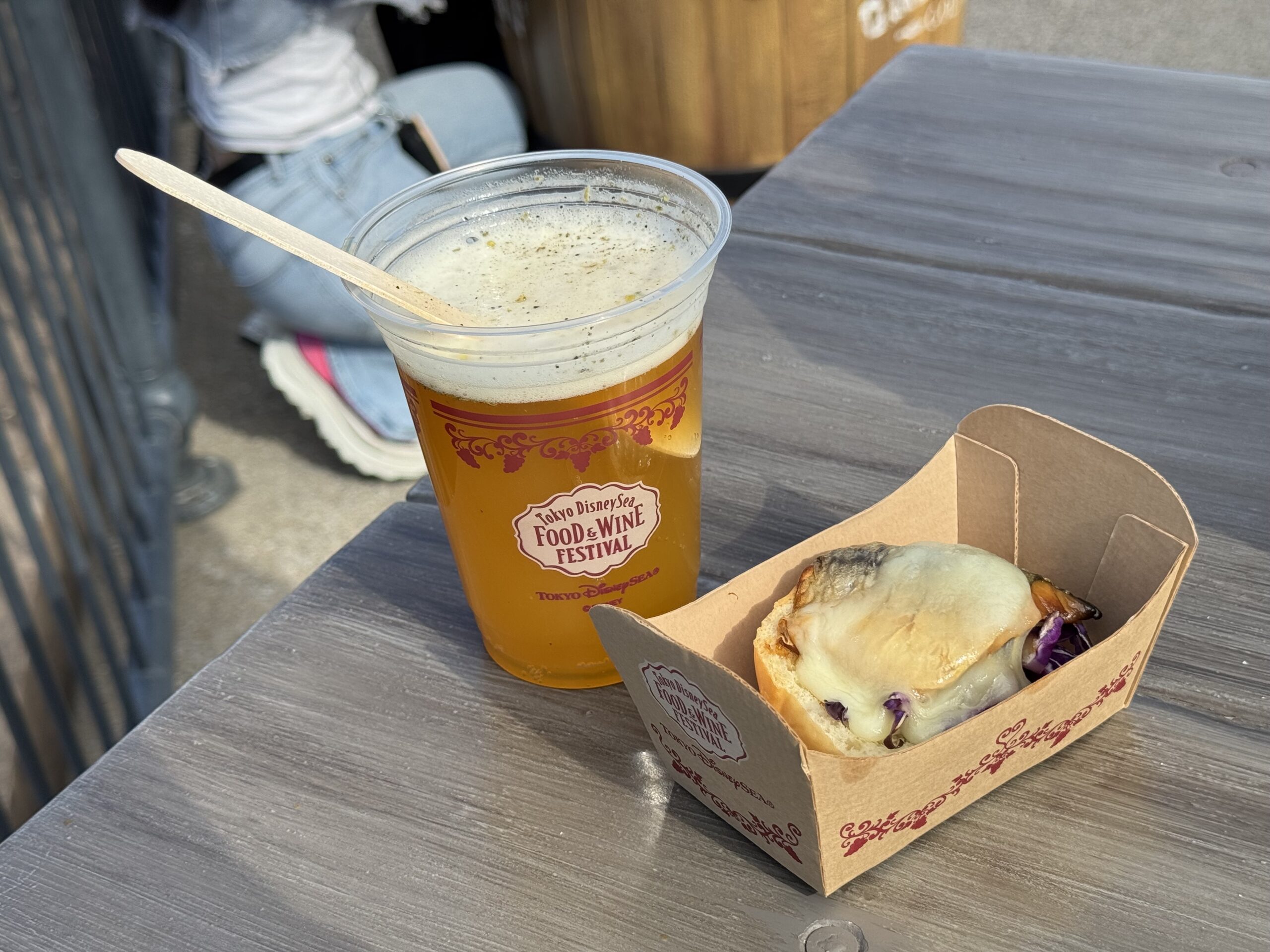At the 2025 Food and Wine Festival at Tokyo DisneySea, a plastic cup of beer and a bread bowl with melted cheese served in a paper tray sit on a wooden table, ready to delight festival-goers.