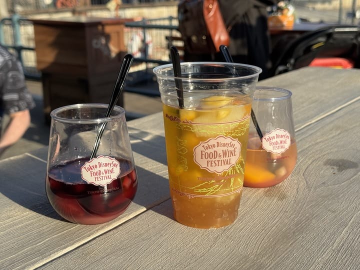 Three festival drinks at the 2025 Tokyo DisneySea Food and Wine Festival, sunlight and crowds in the background.