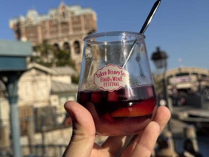 A hand holds sangria labeled "Tokyo DisneySea 2025 Food and Wine Festival" against a blurred park background.