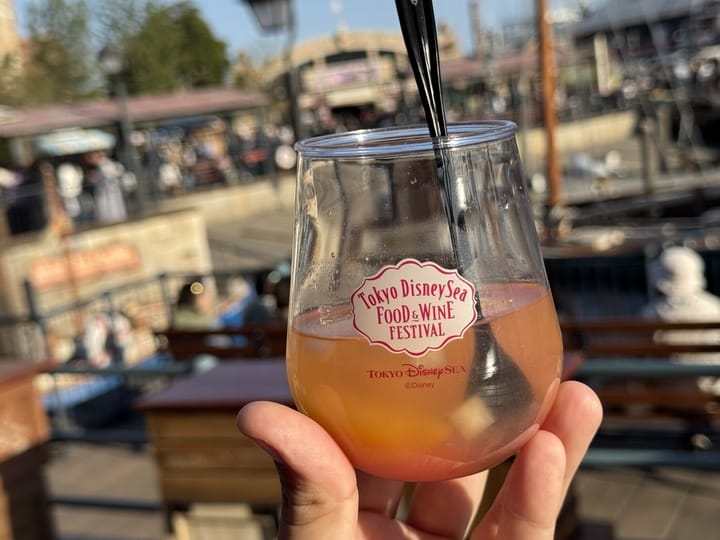 A hand holds a glass of fruity drink at the 2025 Tokyo DisneySea Food and Wine Festival, with a scenic background.
