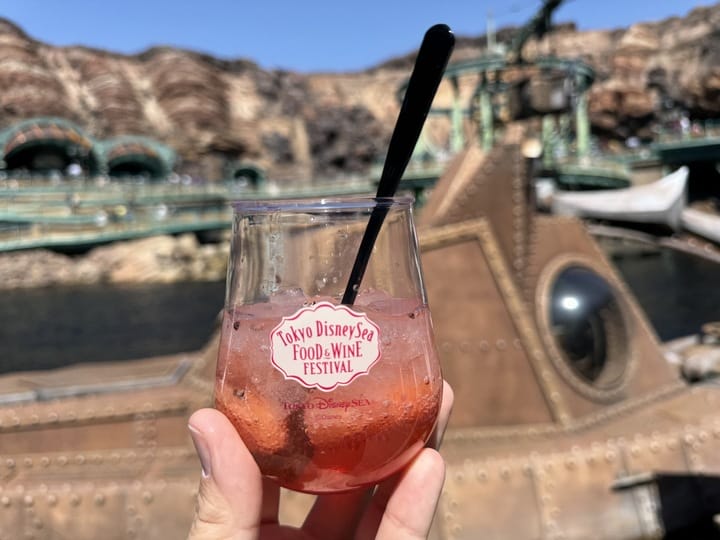 A hand holds a glass of pink drink with ice and a straw, labeled "Tokyo DisneySea Food & Wine Festival 2025," set against a steampunk-themed background.