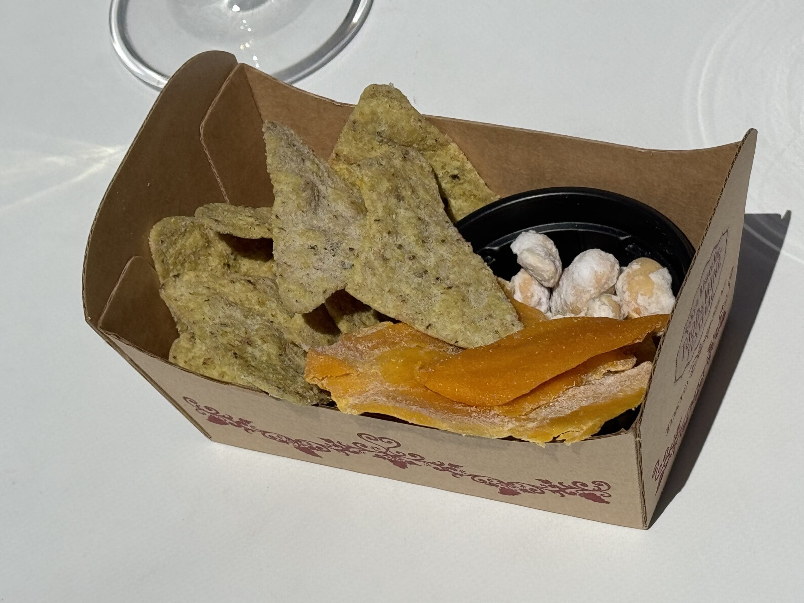 A small box with green tortilla chips, dried mango, and nuts at the 2025 Food and Wine Festival on a white surface.