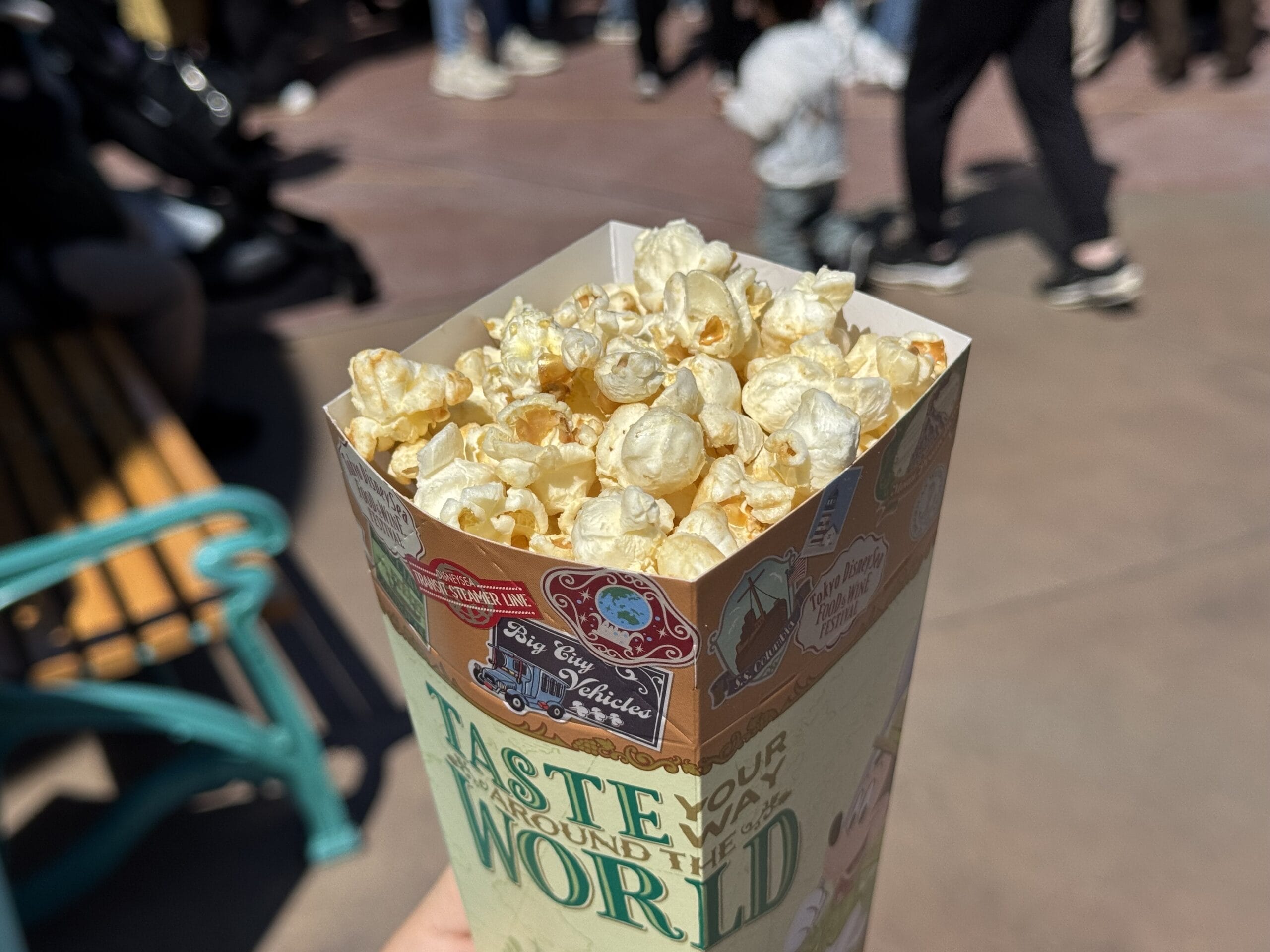 A close-up of a container of popcorn held outdoors at the 2025 Food and Wine Festival in Tokyo DisneySea, with blurred people and a bench visible in the background.