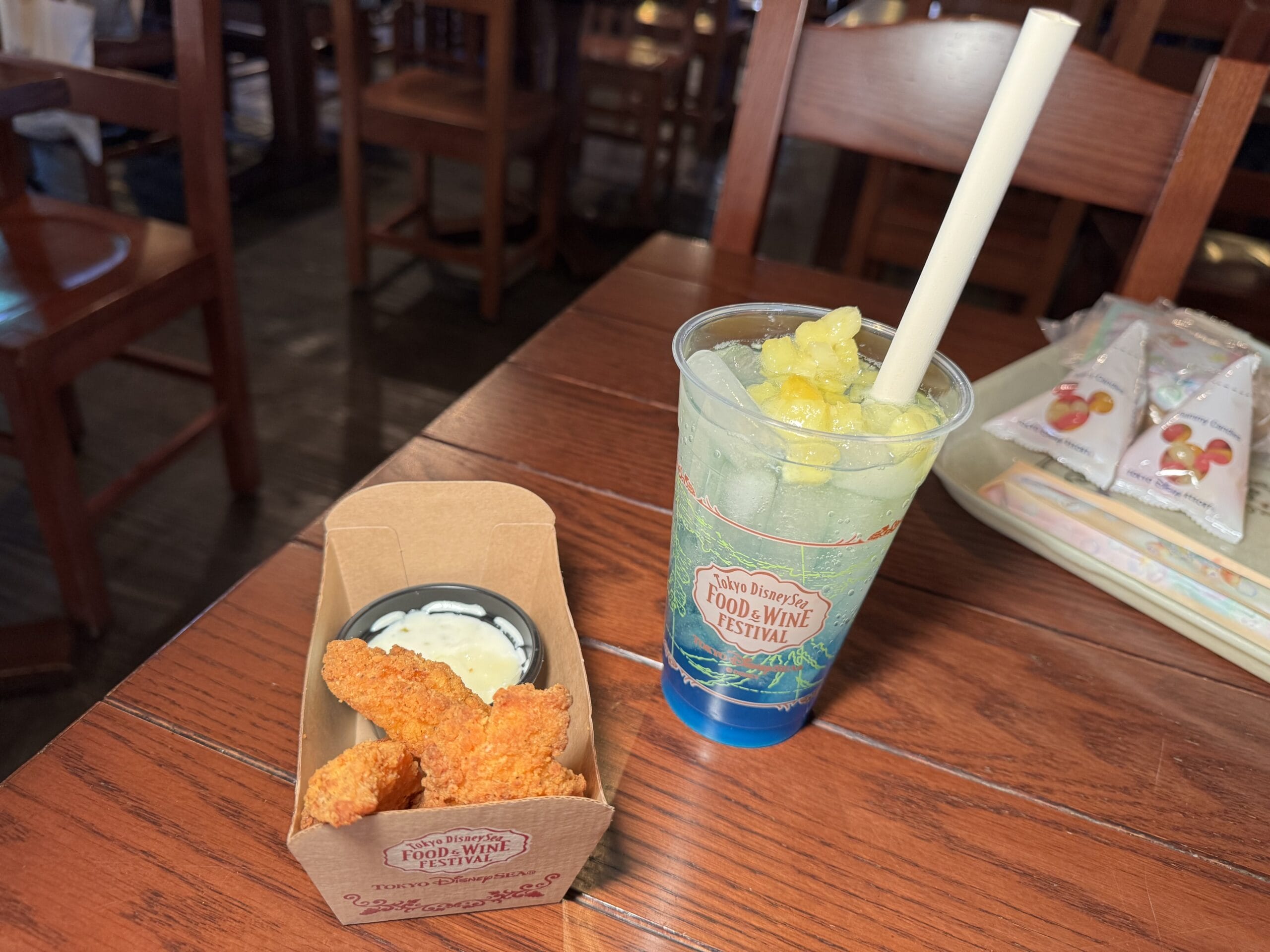 Chicken tenders with dipping sauce in a tray and a blue drink with pineapple pieces in a plastic cup on a wooden table at the 2025 Food and Wine Festival.