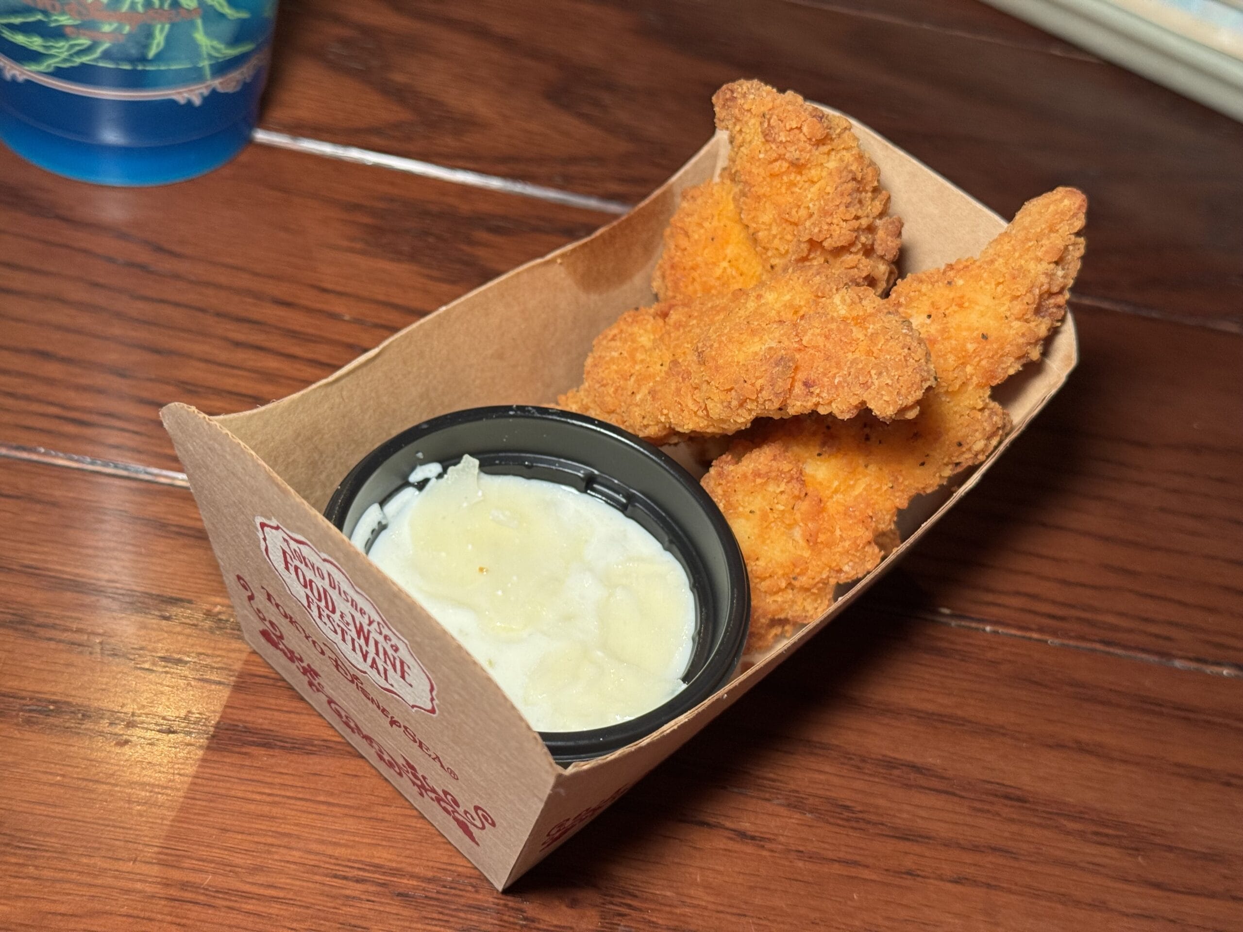 A cardboard container holding breaded chicken tenders with a side of white dipping sauce sits on a wooden table at the 2025 Food and Wine Festival in Tokyo DisneySea.