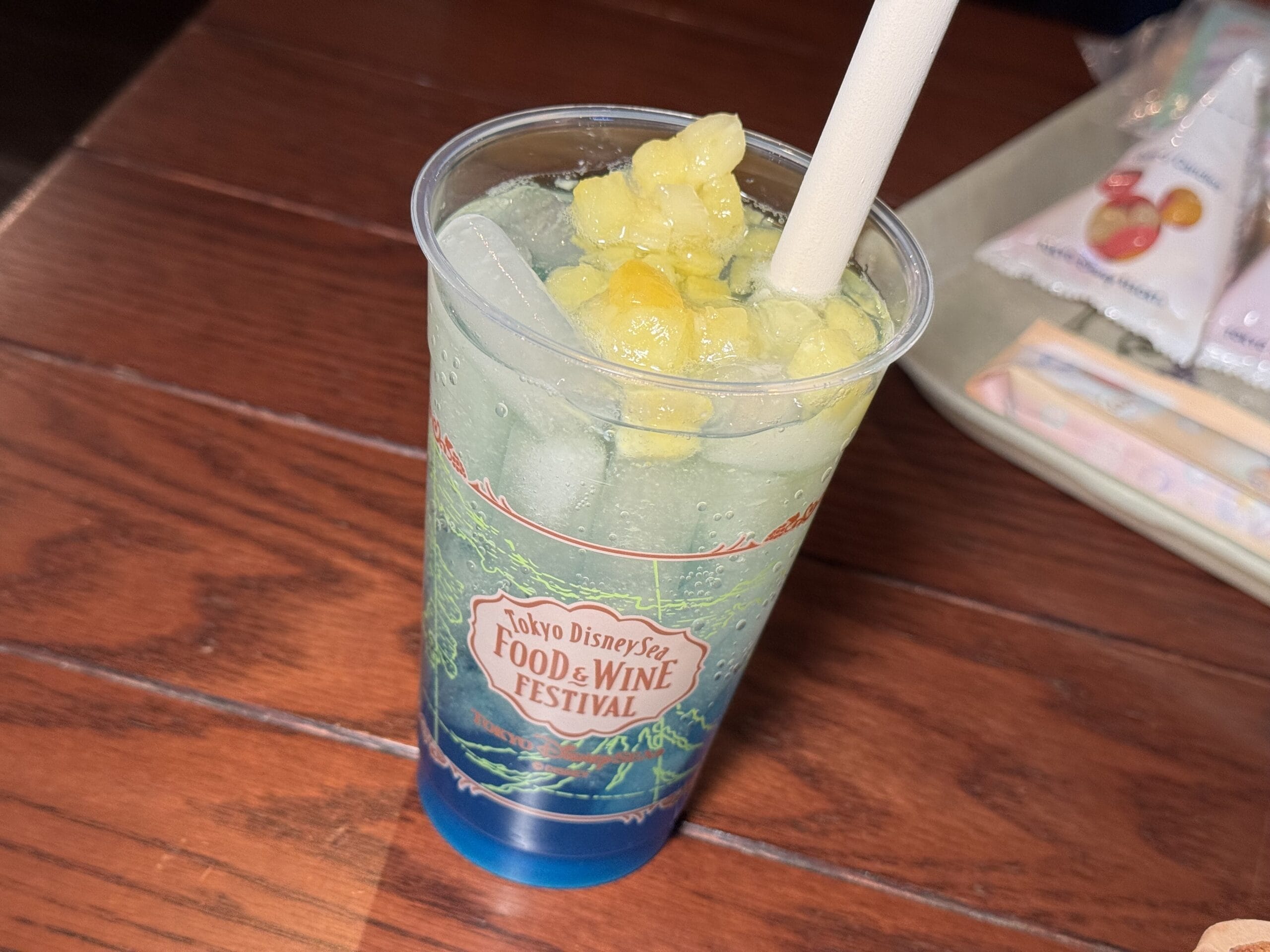 A blue-green iced drink with fruit pieces on top sits in a clear cup labeled "Tokyo DisneySea 2025 Food & Wine Festival," placed on a wooden table.