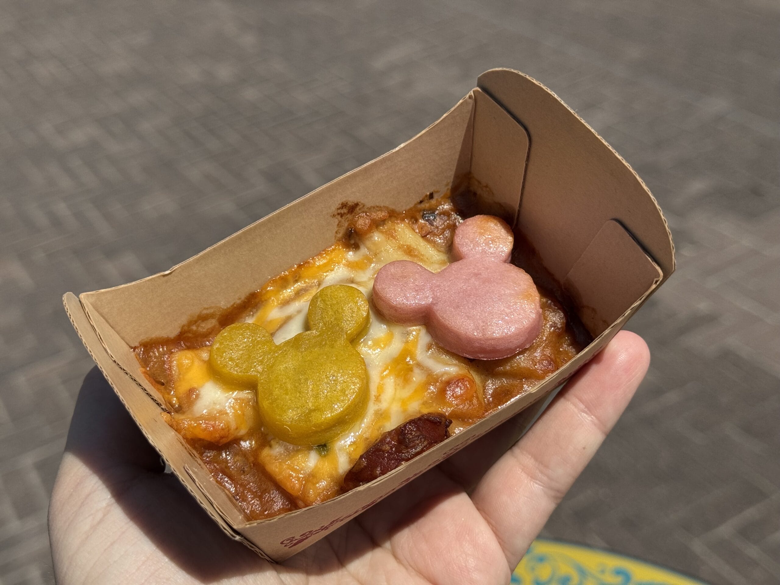 A hand holds a tray of baked pasta topped with melted cheese and two Mickey Mouse-shaped pieces, one yellow and one pink, at the 2025 Food and Wine Festival in Tokyo DisneySea.