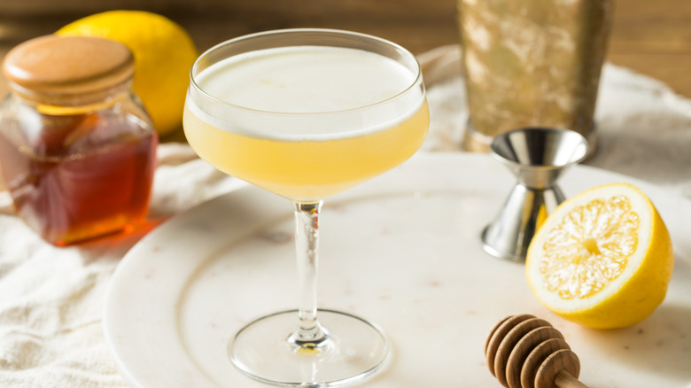 A pale yellow cocktail in a coupe glass with a jar of honey in the background and half of a lemon next to it.