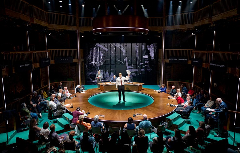 Man stands on a circular stage, surrounded by audience members on chairs.