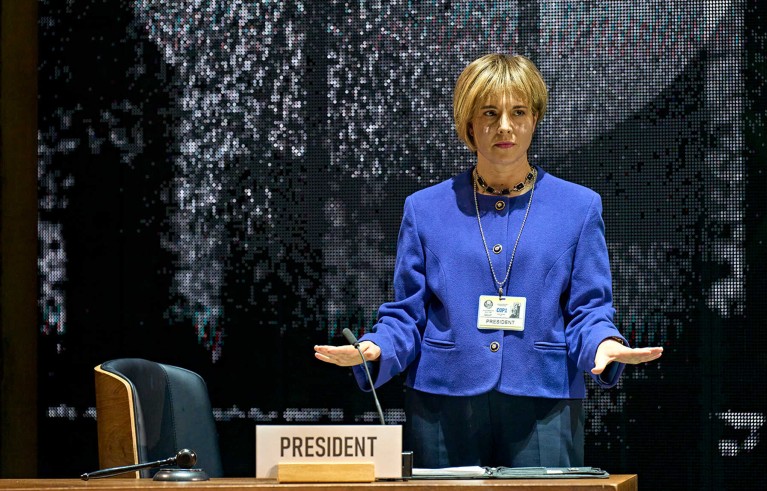 Woman with blonde hair and wearing a blue tailleur stands behind a sign saying ‘president’ in the Kyoto play.