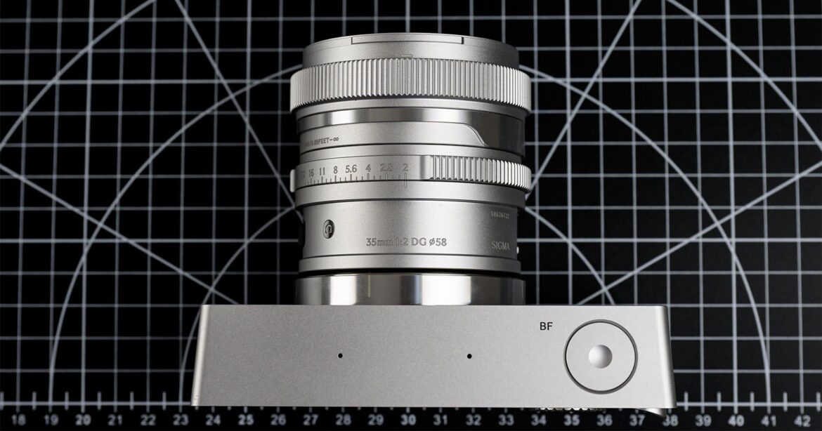 A modern silver camera lens with detailed markings, positioned vertically on a silver surface. The background features a black and white grid pattern with circular and diagonal lines.