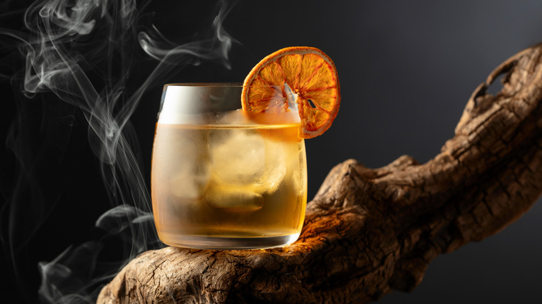 A pale orange cocktail in a rocks glass garnished with an dehydrated orange wheel. Smoke is billowing in the background.