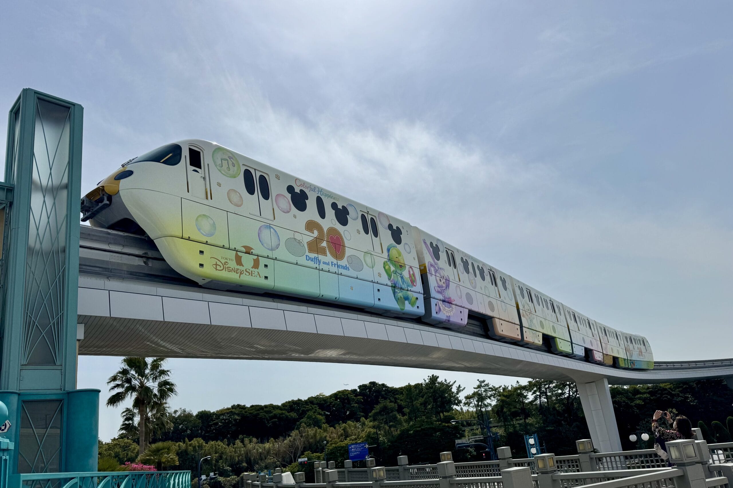 A Tokyo Disney Resort Monorail, decorated with Disney characters and Duffy & Friends 20th anniversary graphics, glides along its elevated track under a clear sky for an enchanting ride.